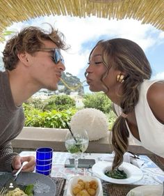 a man and woman are kissing at an outdoor table with food on the plate in front of them