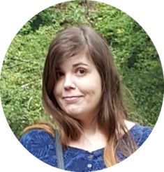 a woman with long brown hair and blue shirt is looking at the camera while standing in front of some trees