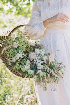 a woman in white dress holding a basket with flowers and greenery on the side