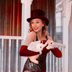 a woman in a red dress and hat holding a white cat while standing on a porch