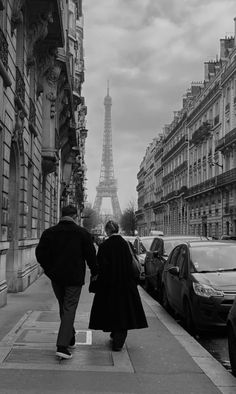 two people walking down the sidewalk in front of cars and eiffel tower behind them