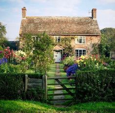 an old brick house surrounded by flowers and greenery with a wooden gate leading to the front door