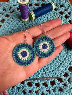 a pair of crocheted earrings is being held by someone's hand next to yarn and thread