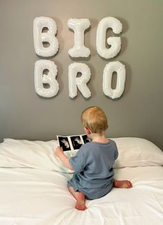 a little boy sitting on top of a bed reading a book in front of balloons that spell out the word big bro