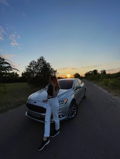 a woman leaning on the hood of a silver car in front of trees at sunset