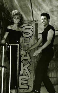 two people standing next to each other in front of a wooden sign with words on it