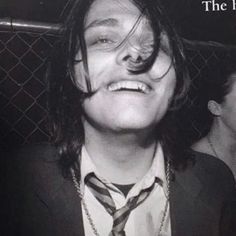 a man with long hair wearing a suit and tie smiling at the camera while standing in front of a chain link fence