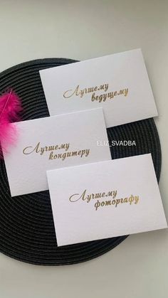 three place cards with gold lettering on them and a pink feather resting on the top