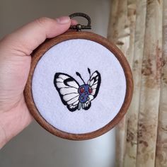 a hand holding a small embroidered butterfly on a piece of wood with white fabric behind it