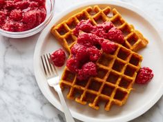 a white plate topped with waffles covered in raspberries next to a bowl of sauce