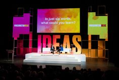 two people are sitting on a stage in front of large screens with words that spell out ideas