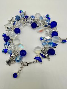 This beautiful charm bracelet features a dazzling combination of blue and white beads, reminiscent of the ocean and sky. The design includes translucent frosted beads, deep royal blue beads, and clear beads, creating a dynamic contrast. The bracelet is adorned with various charms, including a silver star, turtle, butterfly, and evil eye beads, adding a mystical and protective touch. The adjustable chain with a royal blue charm at the end ensures a perfect fit, while the clustering of beads and charms gives it a whimsical, layered look. Evil Eye Beads, Blue Charm, Cluster Bracelets, Deep Royal Blue, Clear Beads, Eye Beads, White Beads, Silver Stars, Blue Beads