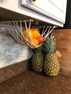 pineapples, oranges and apples are in a basket on the kitchen counter