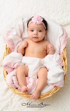 a baby is sitting in a wicker basket on a white blanket and wearing a pink bow