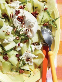 a yellow plate topped with cucumber, cheese and pecans next to a spoon