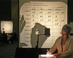 a woman writing on a piece of paper in front of a projection screen with the shadow of a person behind her