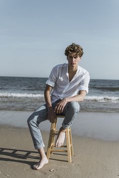 a young man sitting on top of a wooden stool next to the ocean with his legs crossed