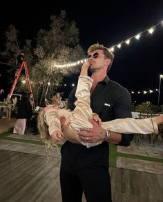 a man holding a woman in his arms at an outdoor event with string lights on the background