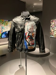 a black leather jacket with graffiti on it is sitting on a stand in front of two paintings