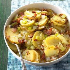 a white bowl filled with potatoes and bacon on top of a blue cloth next to a spoon