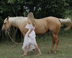 a woman in white dress walking next to a brown horse