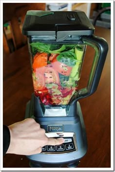 a blender filled with lots of vegetables on top of a wooden table next to a person's hand