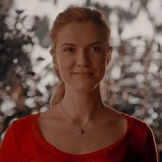 a woman with blonde hair wearing a red shirt and necklace in front of a tree