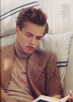 a young man sitting on top of a bed wearing a brown suit and turtle neck sweater