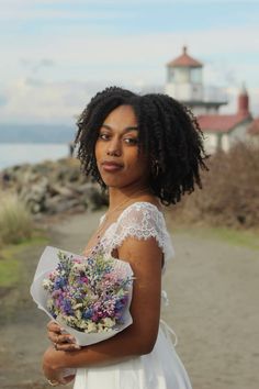 a woman holding a bouquet of flowers in front of a light house on the beach