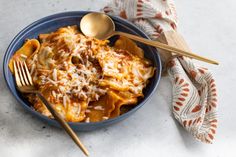 a blue bowl filled with pasta and cheese on top of a white tablecloth next to two gold spoons