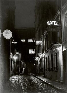 a black and white photo of people walking down a street at night with hotel signs on the buildings