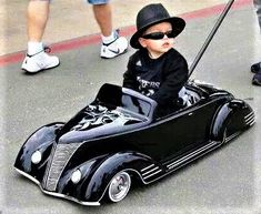 a small child in a black toy car on the street with people standing around it