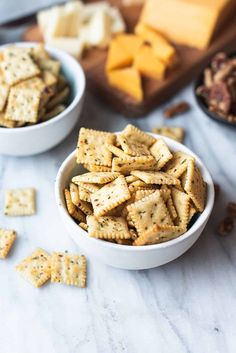 two white bowls filled with crackers next to cheese and nuts on a marble counter