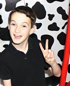 a young boy making the peace sign in front of a cow print wall with his fingers up