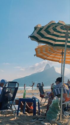 people sitting under umbrellas at the beach on a sunny day with mountains in the background