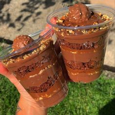 two desserts in plastic cups are being held by someone's hand with grass behind them