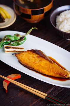 a white plate topped with fish next to rice and chopsticks on a wooden table