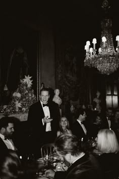 a man in a tuxedo standing at a dinner table with people seated around it