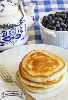 pancakes and blueberries on a plate with a teapot, fork and knife next to it