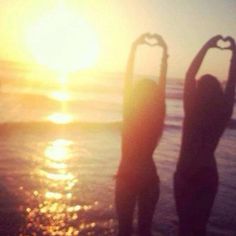 two people standing on the beach making a heart shape with their hands in front of the sun