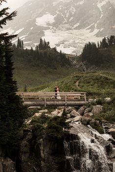 a bride and groom standing on a bridge over a waterfall in front of a mountain