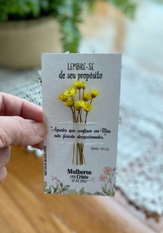 a person holding up a small card with yellow flowers on it in front of a potted plant