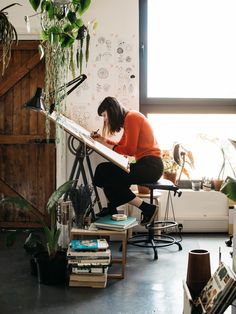 a woman sitting at a desk in front of a window with plants and books on it