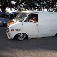 a white van parked in a parking lot next to other cars and trees with one man sitting in the driver's seat