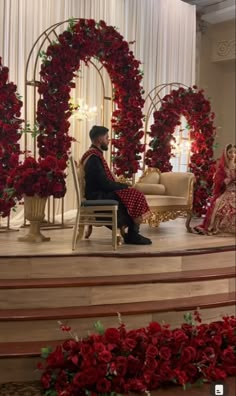 a man and woman sitting on stage with red flowers in front of them at a wedding