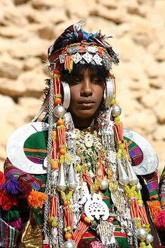 a woman in an elaborate headdress stands near some rocks and looks at the camera