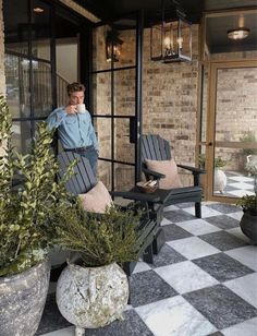 a man is standing on the porch next to two chairs and plants in front of him