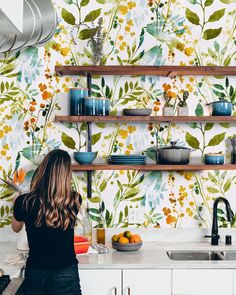 a woman standing in front of a kitchen counter next to a wall with flowers on it