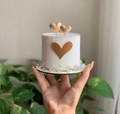 a hand holding a white cake with gold hearts on top and greenery around it