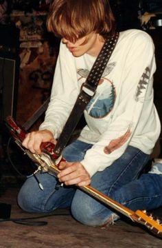 a young man sitting on the floor with his guitar strap around his neck and looking down